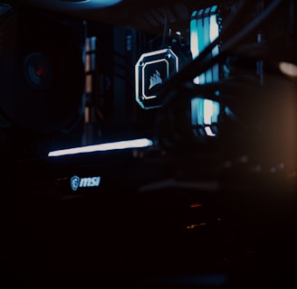 A close-up view of a computer gaming rig illuminated with blue LED lights. The interior shows components like cooling fans at the top, a glowing logo on a central component, and an MSI branded part at the bottom. The scene is dark, emphasizing the glow of the lights.