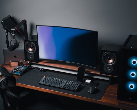 A modern gaming and streaming setup with a curved monitor placed centrally on a wooden desk. Surrounding the monitor are powerful speakers and a high-end PC tower with glowing fans. Various peripherals, including a mechanical keyboard, ergonomic mouse, and audio mixer, are neatly arranged. Overhead, a professional microphone and stand are ready for use, possibly for gaming commentary or podcasting.